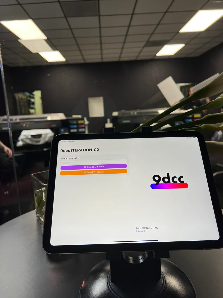 A tablet showing the 9dcc ITERATION-02 minting interface, with garment printers in the background.
