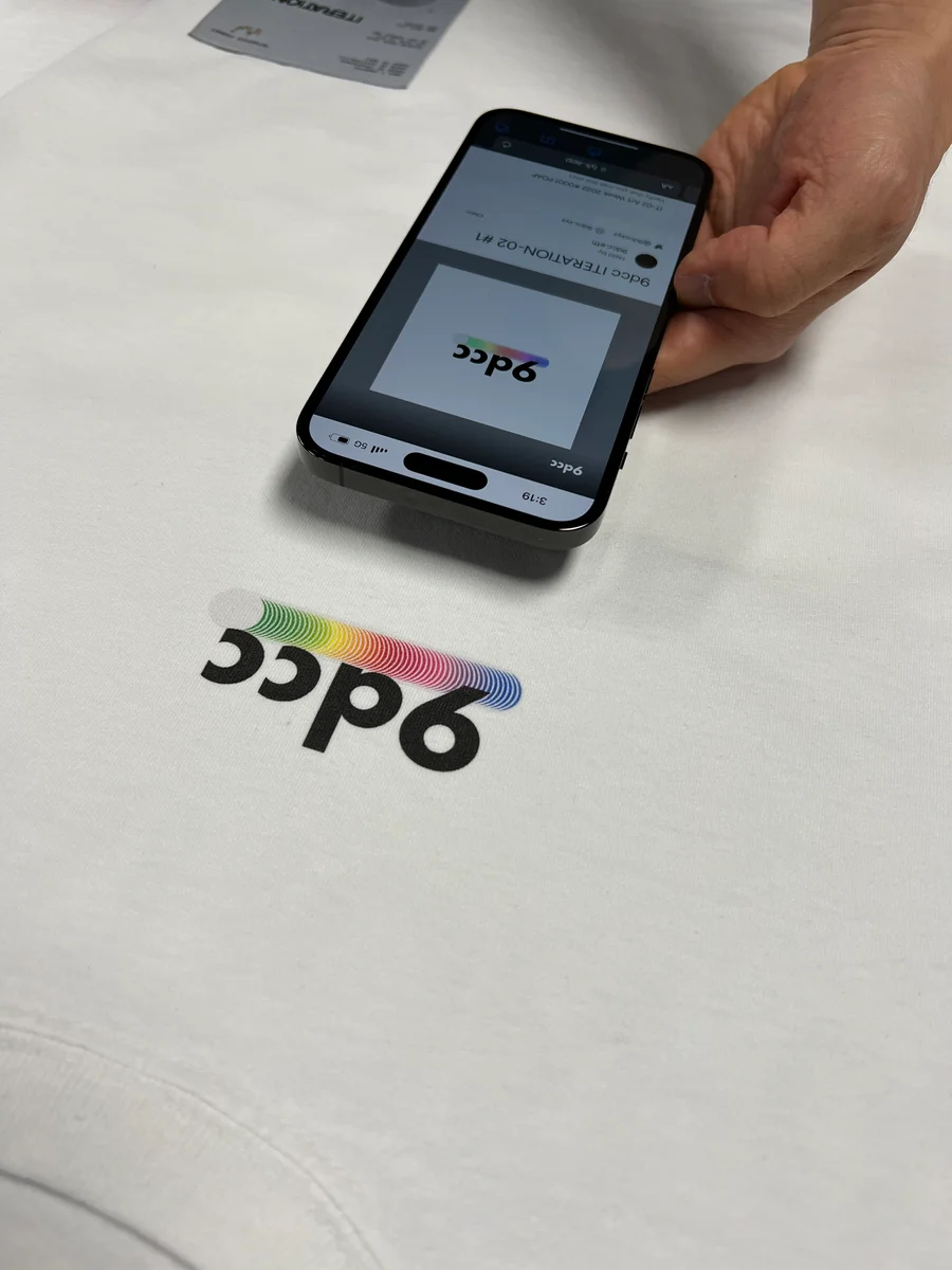 A phone displaying a 9dcc ITERATION-02 artwork rests beside its matching rainbow print on a white shirt.