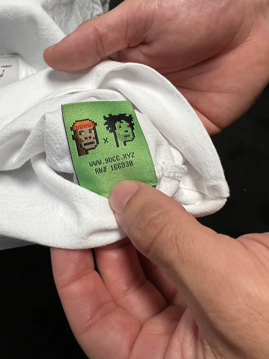 Hands holding open a white shirt to show a green woven 9dcc label with two pixel-art portraits.