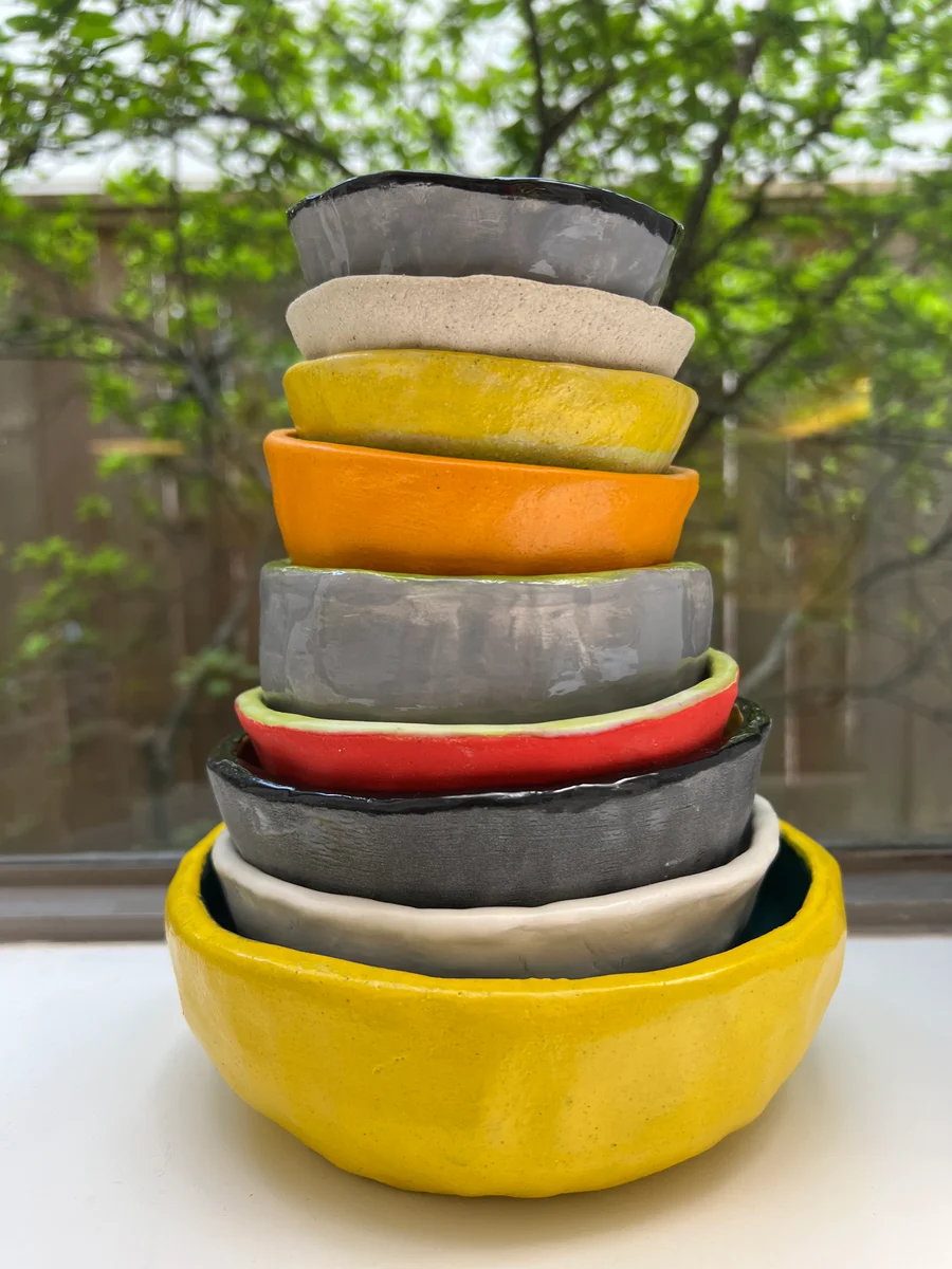 An uneven stack of colorful handmade bowls beside a window, showing their different rims, sizes, and surfaces.