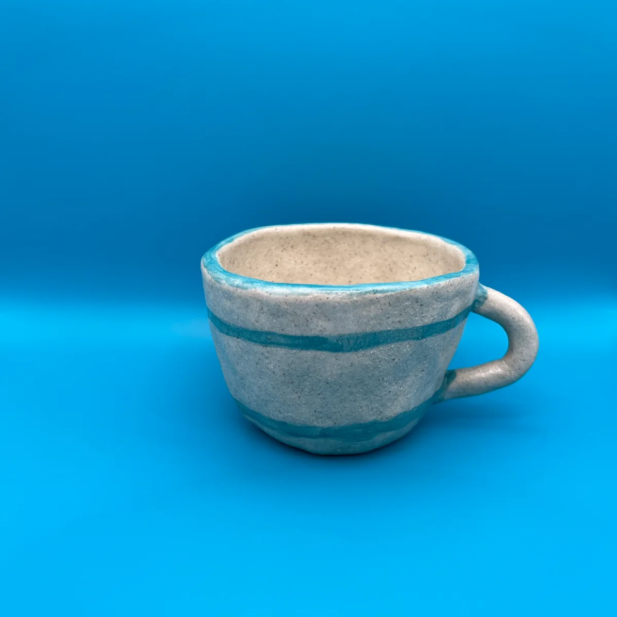 A pale speckled ceramic cup with turquoise stripes and a handle, photographed against a bright blue background.