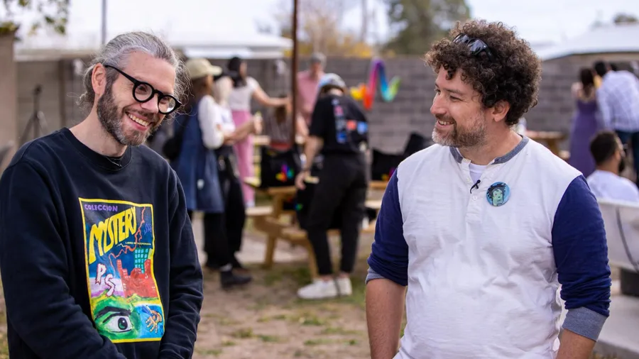 Alexis André on the left and Erick Calderon on the right, smiling together outdoors with people gathered behind them.