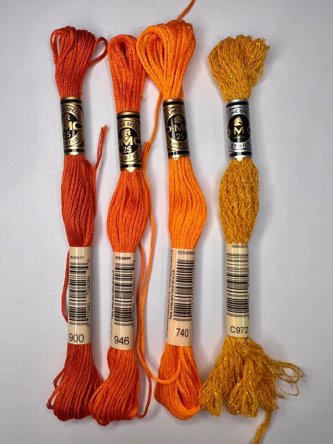 Four warm-colored DMC skeins labeled 900, 946, 740, and C972, laid side by side on a pale background.