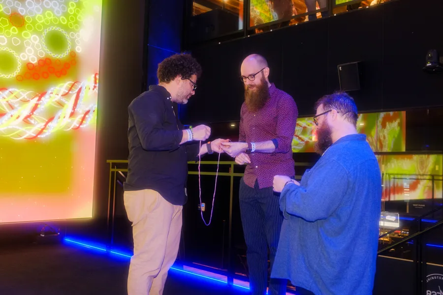 Erick and two attendees holding threads and making bracelets, with colorful Friendship Bracelets artworks on the screens behind them.