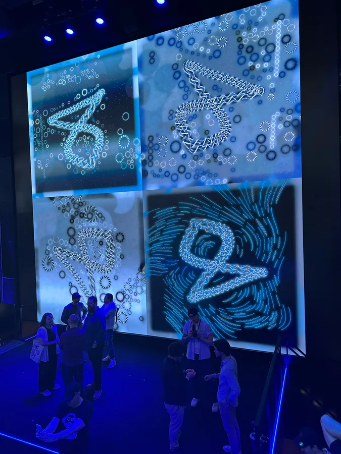 Four blue-toned Friendship Bracelets artworks fill a towering screen, with people gathered below it.