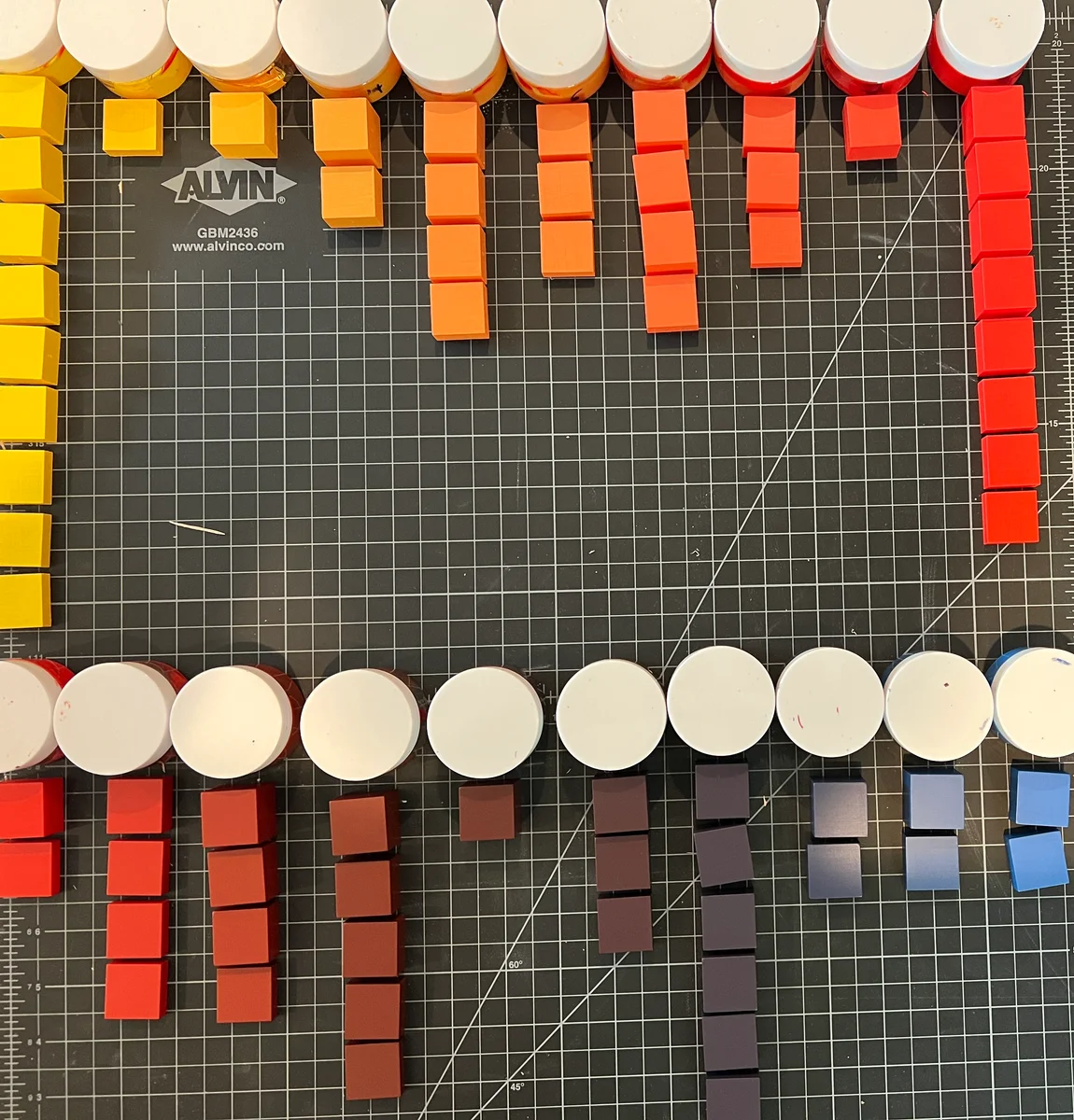 Painted blocks arranged in rows beside paint jars on a gridded cutting mat, shifting from yellows and oranges to reds, purples, and blues.