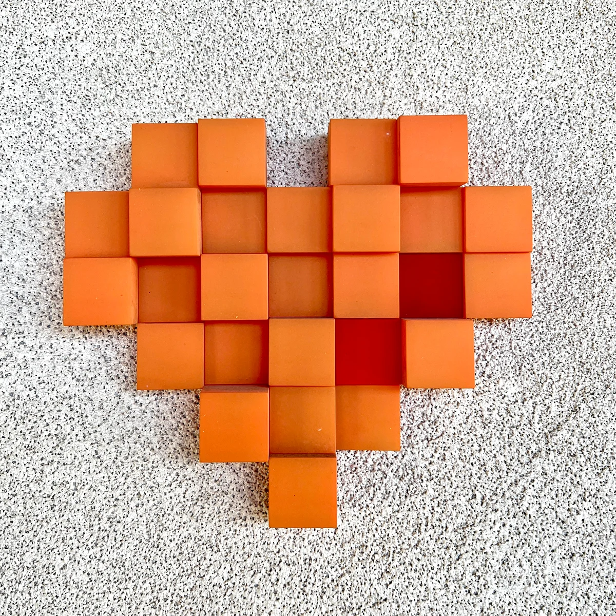 An orange block heart on a textured pale wall, with shadows revealing the different block heights.