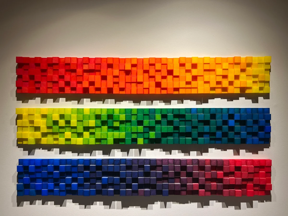 Three long horizontal block sculptures mounted on a wall, with varying depths and colors progressing through the spectrum.