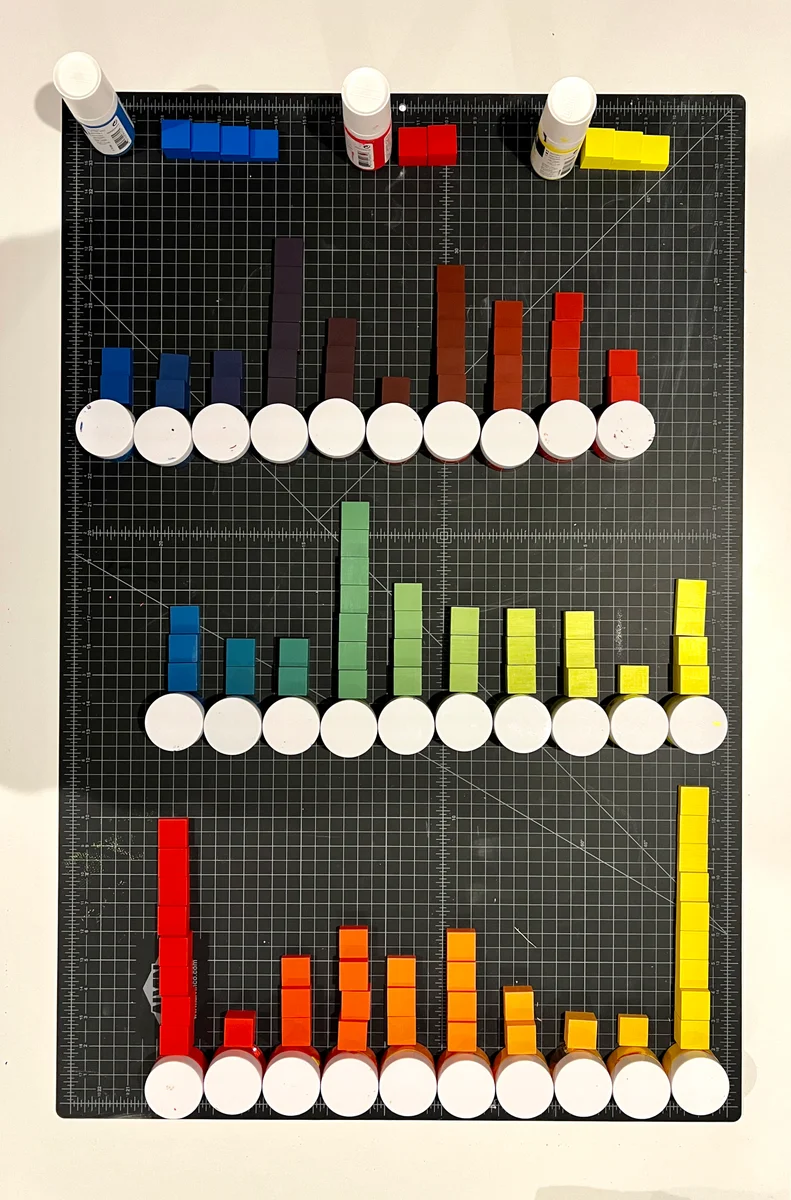 An overhead view of blocks grouped by paint color across a cutting mat, with paint jars and blue, red, and yellow paint bottles.