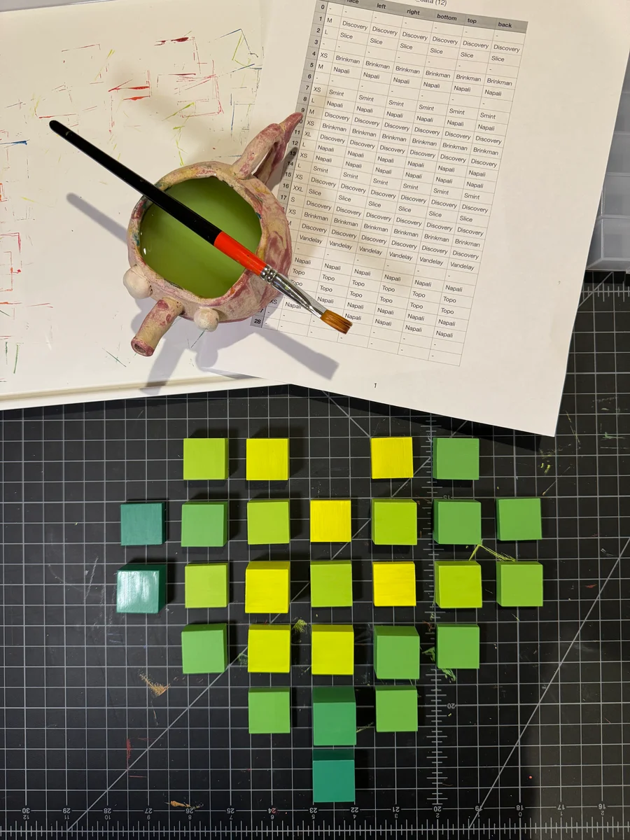 Green and yellow blocks laid out in a heart shape on a cutting mat, beside a printed block-by-block color sheet and a paintbrush.