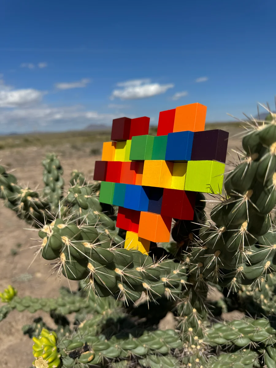 The same rainbow-colored block heart among cactus pads in Marfa, with desert and blue sky behind it.