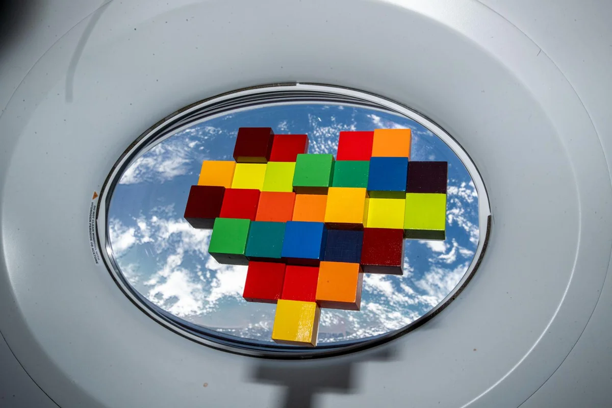 A hand-painted block heart in front of a spacecraft window, with Earth's blue surface and white clouds below.