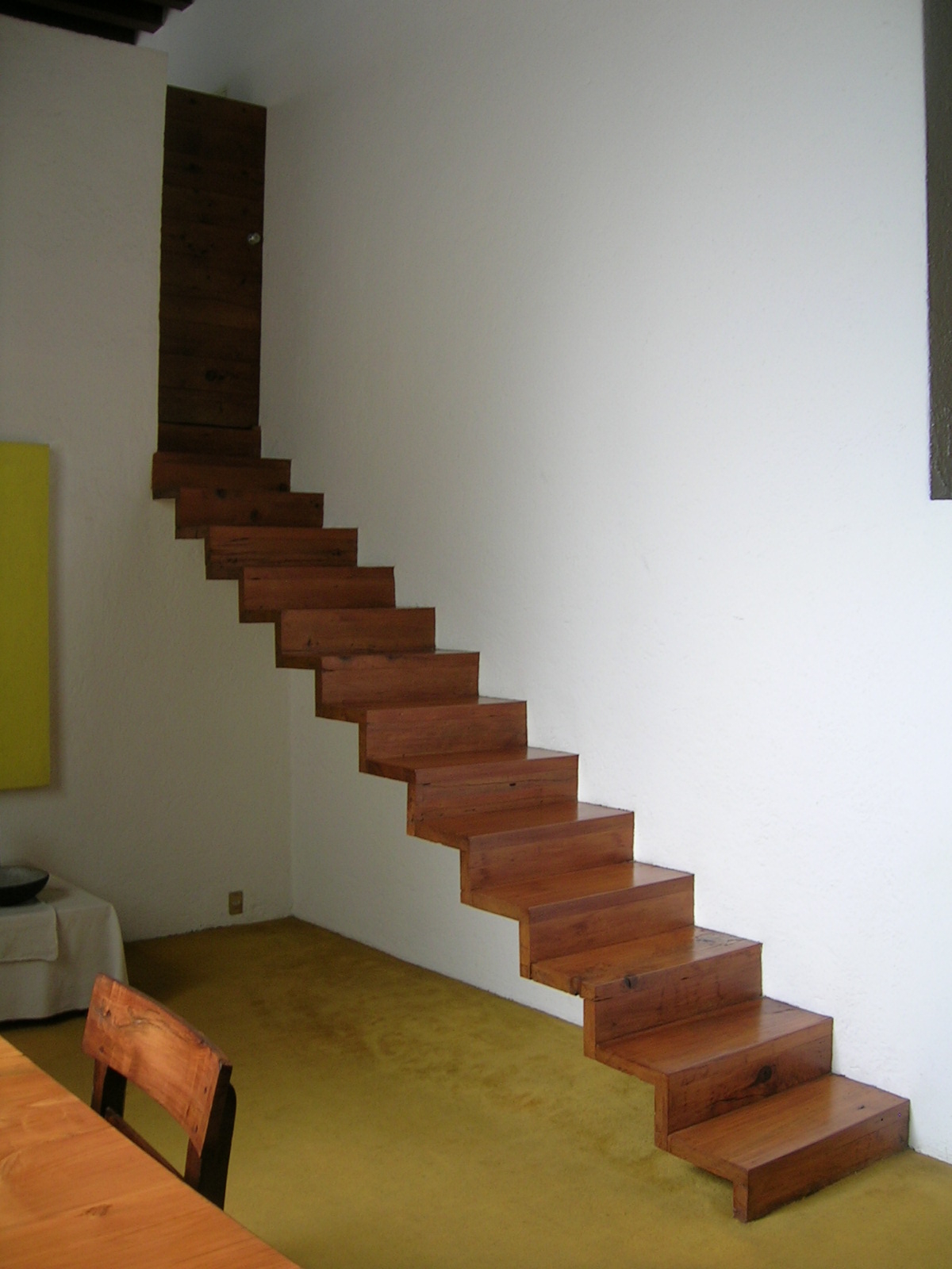 A wooden staircase without a railing rises along a white wall to a narrow doorway at Luis Barragán House and Studio.