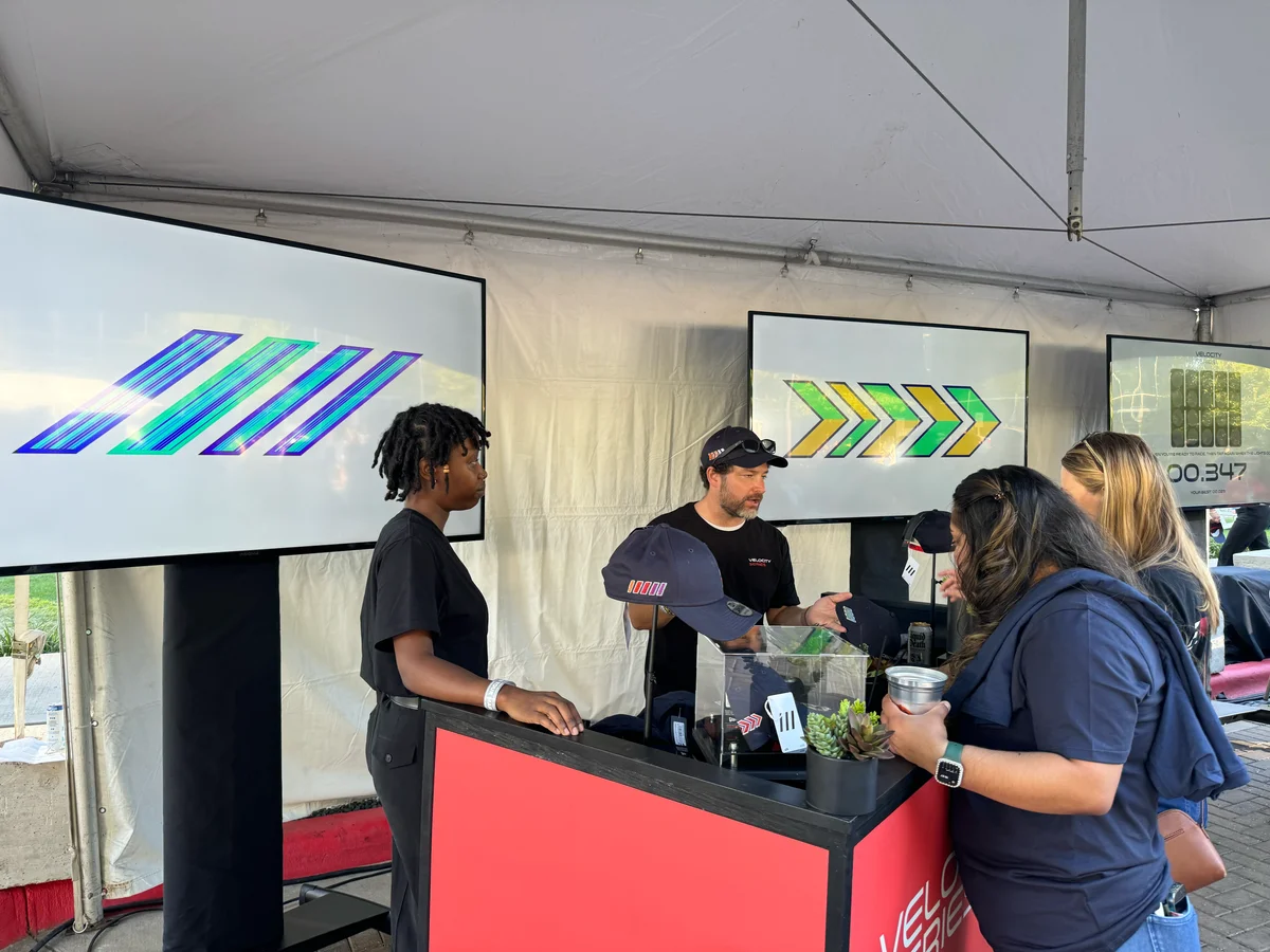 Visitors talking at a booth displaying embroidered caps, with large screens showing slash and chevron artworks behind the counter.