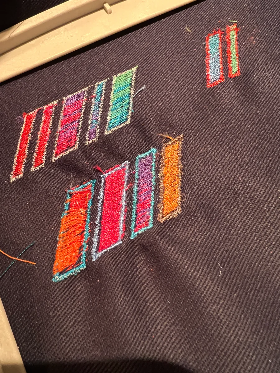 Several groups of embroidered slashes on dark fabric held in a hoop, with multicolored fills and contrasting borders.