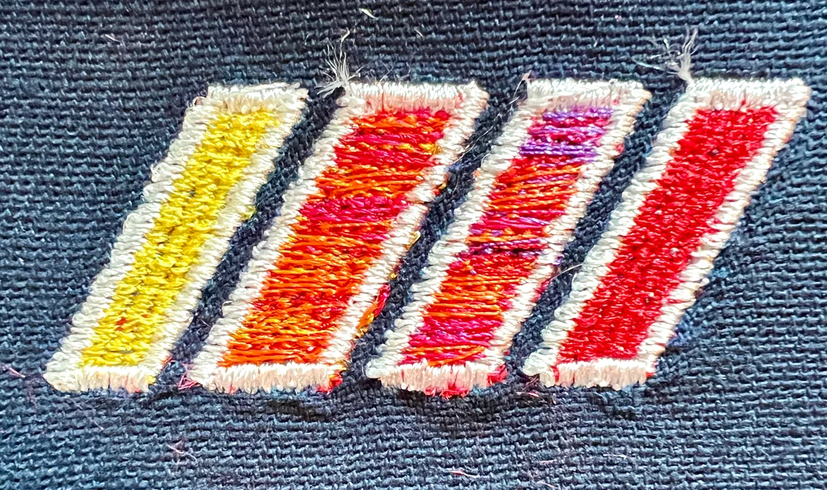 A close-up of four embroidered slashes on dark fabric, with white borders and yellow, orange, pink, purple, and red thread inside.