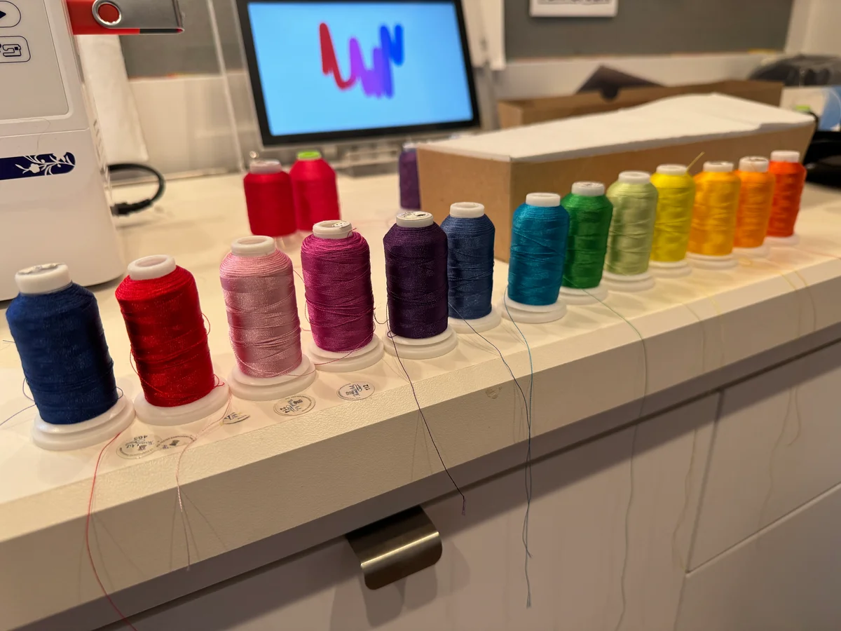 Spools of embroidery thread arranged across a worktop beside a sewing machine, with a colorful Squiggle on a screen behind them.