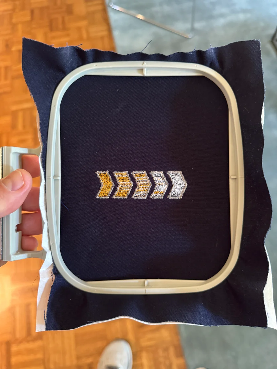 A hand holding an embroidery hoop with five small yellow-and-white chevrons stitched on dark fabric.