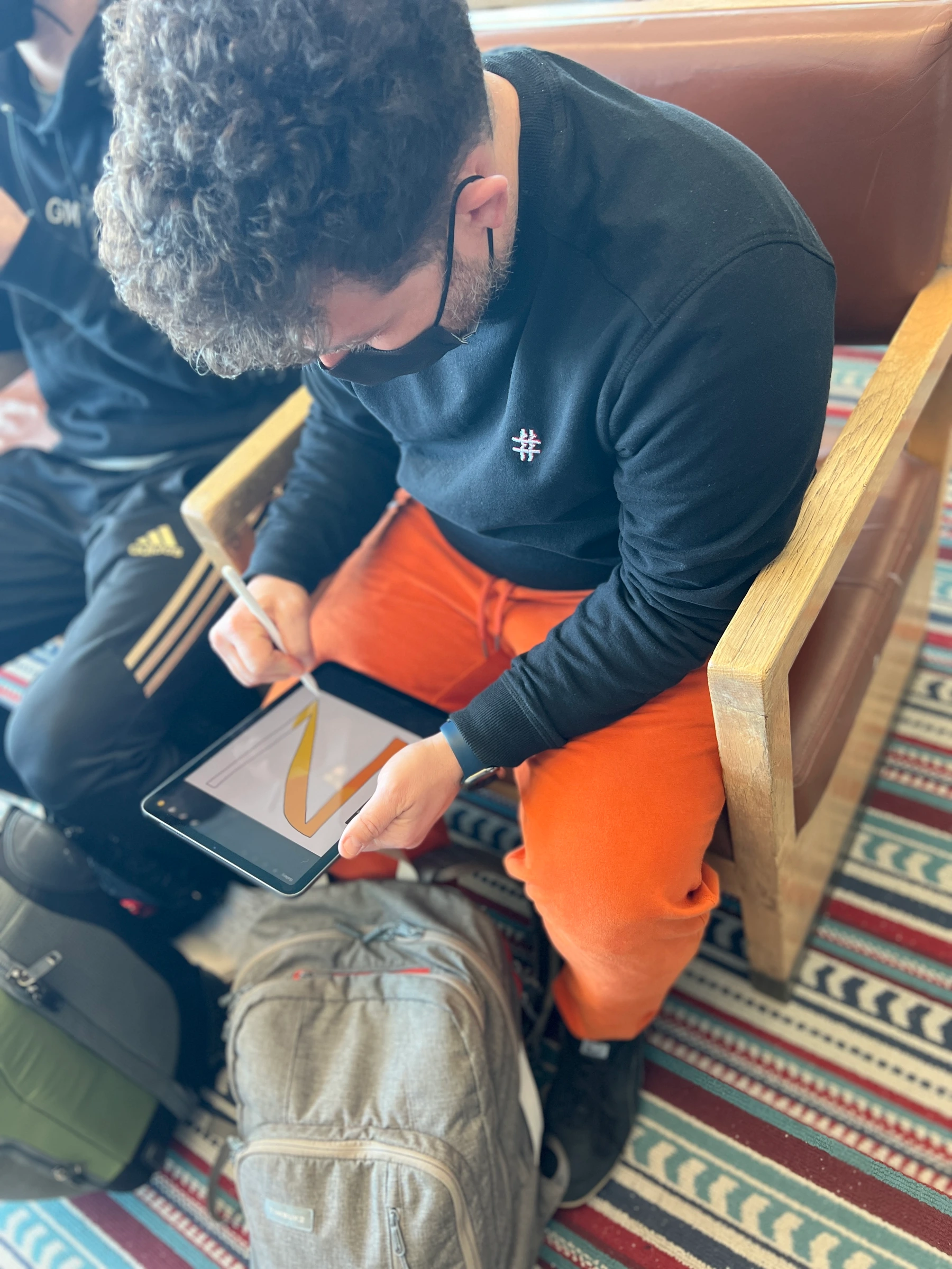 Drawing color into a V on a tablet with a stylus, while seated with the tablet resting on orange trousers.