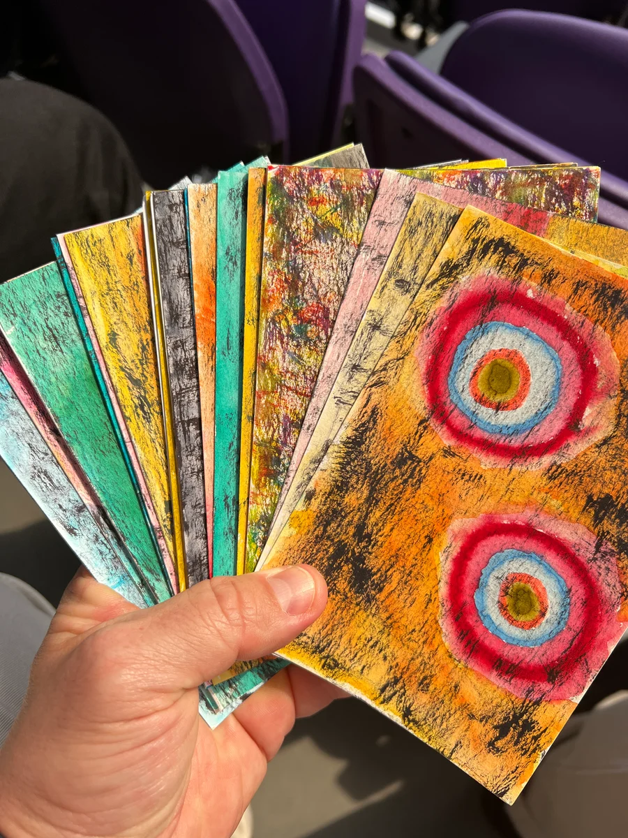 A hand holding a fan of colorful watercolor postcards, with textured surfaces and concentric circles on the front card.